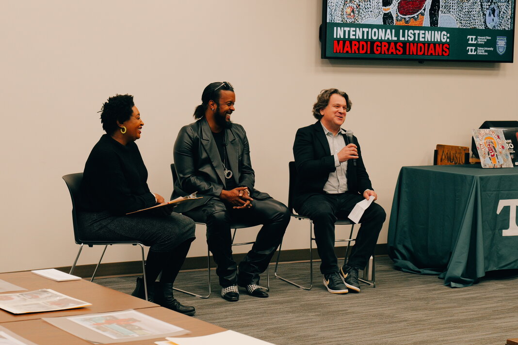 Three speaker at a lecture sit at the front of a room. One holds a microphone in their hand.