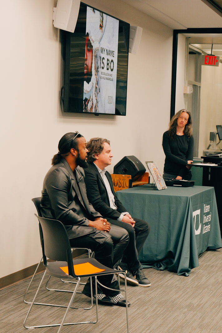 Two speakers sit in chairs at the fron tof a room. One is speaking. In the background the lecture moderator stands behind a table.