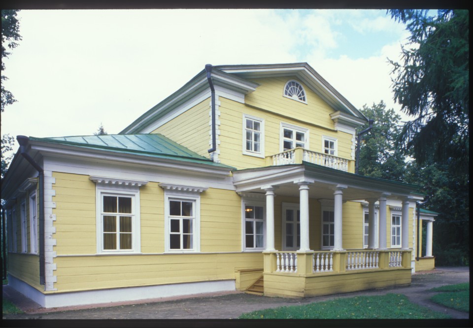 A large, two-story yellow historic house with white columns, a covered front porch, green metal roof, and surrounding trees under a bright sky.