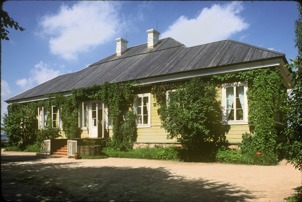 A historic yellow country house with a dark roof, white-trimmed windows, and lush green vines climbing across its façade, viewed from a gravel drive on a sunny day.